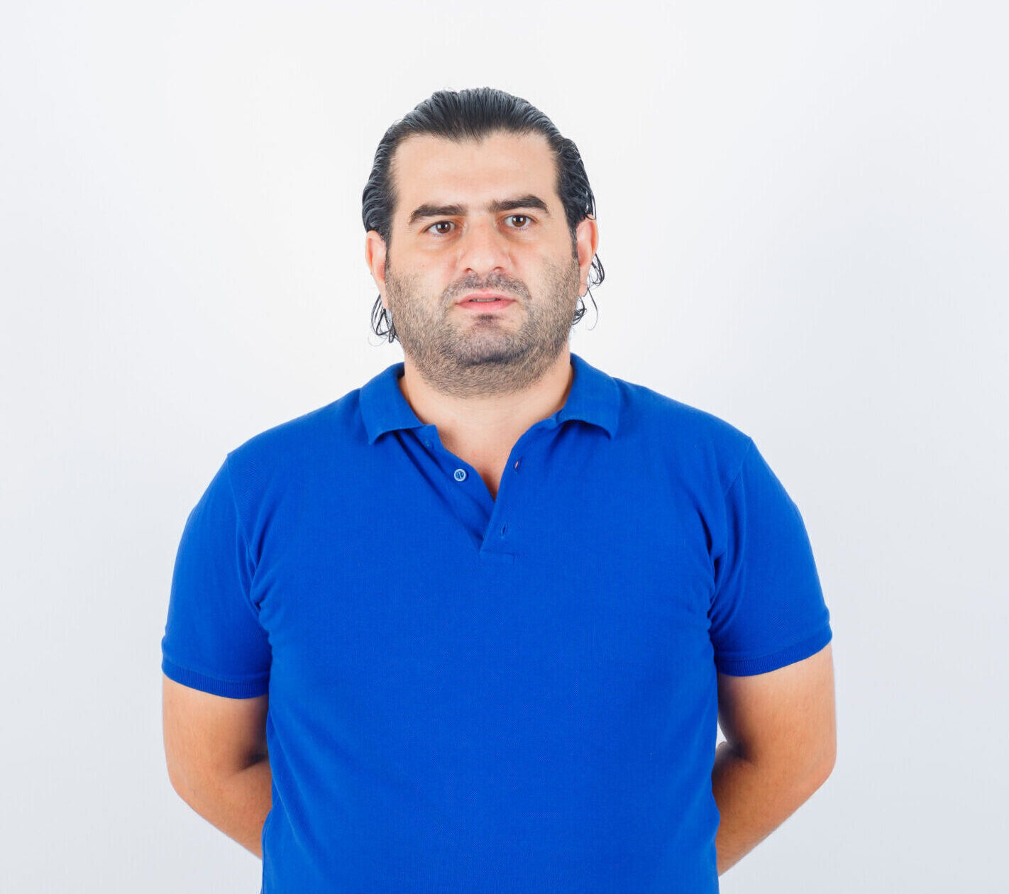 middle aged man in blue t-shirt holding hands behind back and looking confident , front view.